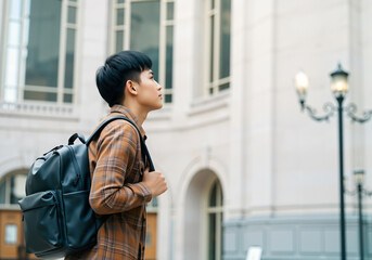 Asian student walking in urban setting near university carrying backpack looking up
