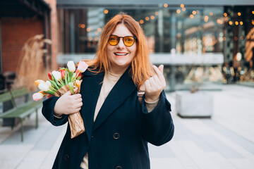 Fototapeta premium Female hand showing finger heart or mini heart gesture with the index finger and thumb come together like a snap outdoors. 30s Woman in sunglasses on urban background. High quality photo.