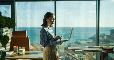 Young professional woman using a laptop in a bright office with a panoramic ocean view, working while standing near large windows in a modern workspace