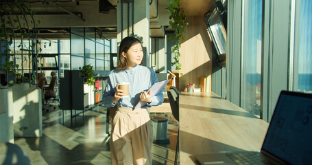 A young professional woman holding a coffee cup and clipboard in a modern office environment with...