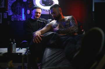 Fototapeta premium bearded tattoo artist working at his studio tattooing sleeve on the arm of his male client. Man getting tattooed by professional tattooist