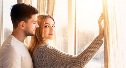 A couple stands together by bright, large windows, adjusting light curtains. The warm sunlight creates a romantic atmosphere, enhancing their intimate moment and cozy setting.