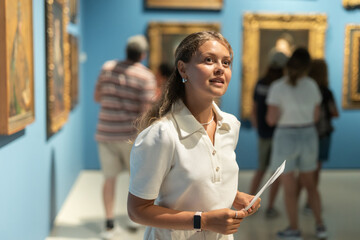 Museum female visitor with paper in hands walk through exhibition halls museum. Girl with guidebook...