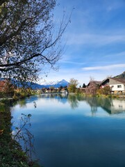Fototapeta premium A serene river flows through a picturesque town, framed by greenery and snow-capped mountains under a vibrant blue sky