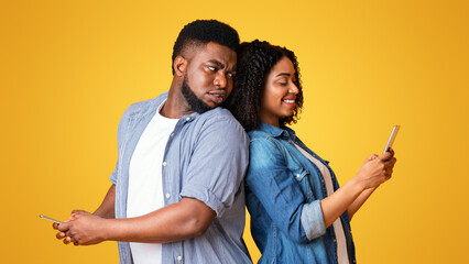 A young couple stands back to back, each engrossed in their smartphone screens. They display...