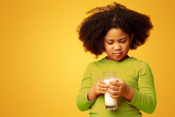 Grumpy little girl refusing to drink milk, looking at glass with disgust, standing over yellow...