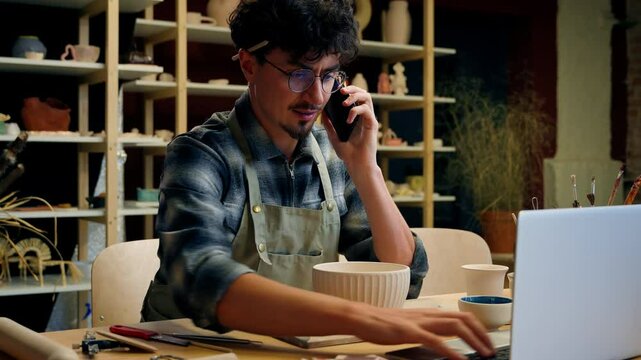 Caucasian man guy potter salesman consult sell ceramics online store small business owner talk mobile phone conversation male sculptor artist typing laptop in pottery workshop craft handmade commerce