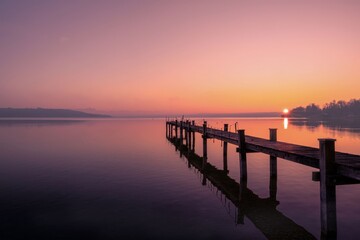 Fototapeta premium A photograph taken from a distance about a serene sunset over a calm lake with a wooden pier extending into the distance. the sky is a gradient of orange and purple hues, with the sun 