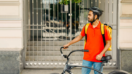 Search for clients address and delivery parcels in city. Serious young arabian guy courier in orange t-shirt and protective helmet, with big backpack with bike stay near customer home, outdoor