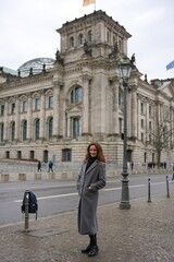 Fototapeta premium A stylish woman in a long gray coat smiles while posing in front of the historic Reichstag building in Berlin, with German and EU flags waving above.