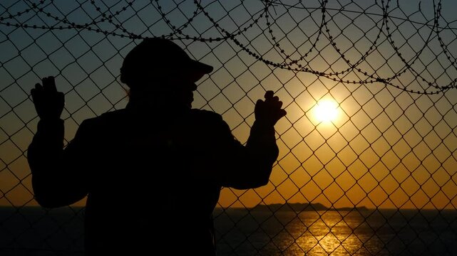 Thirst for a new life. A silhouette of a refugee woman at a barbed wire border looks at the sunset with hope for a new life