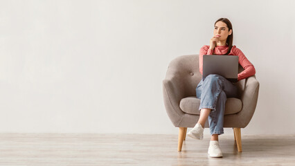 A young brunette woman sits thoughtfully in a cozy armchair, using her laptop in a simple,...