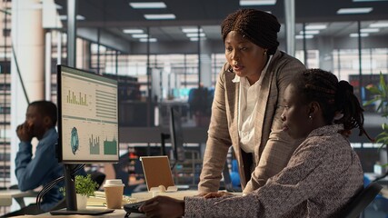 Female employee and manager gathered around a workstation discussing objectives and strategizing...