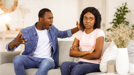 Relationships problems and crisis concept. Angry black man and woman sitting on couch at home, having quarrel, copy space. Furious african american guy yelling at his upset girlfriend