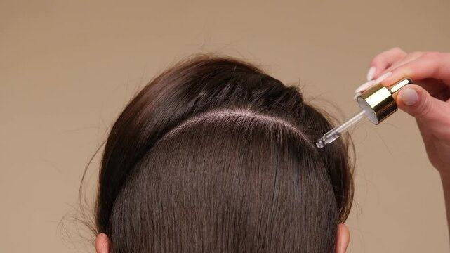 Beautiful woman with brown healthy hair applying oil or serum to roots on beige background. Hair beauty