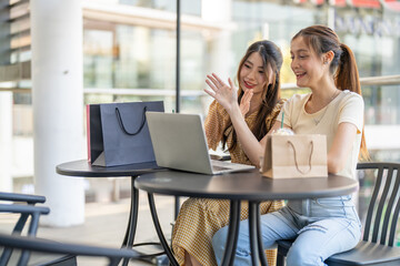 Two diverse women smiling and talking while use laptop for online shopping at cafe, searching for deals and discounts with shopping bags on table, concept of e-commerce, retail, digital lifestyle
