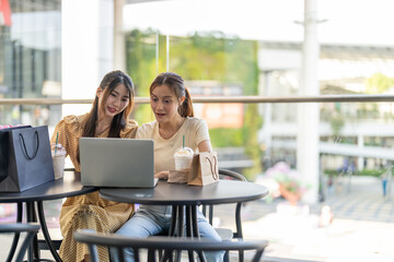 Two diverse women smiling and talking while use laptop for online shopping at cafe, searching for deals and discounts with shopping bags on table, concept of e-commerce, retail, digital lifestyle