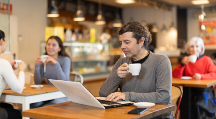 Middle-aged man sitting at table, using laptop and enjoing cup of tea in coffee house. Modern lifestyle concept