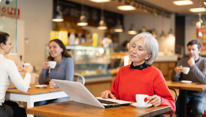 Elderly woman sitting at table, using laptop and enjoing cup of tea in coffee house