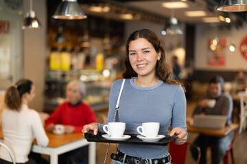 Girl visitor in self-service cafeteria carries tray with two cups of coffee. Customer has purchased freshly brewed coffee for herself and companion