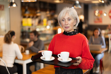 Senior woman visitor in self-service cafeteria carries tray with two cups of coffee. Customer has purchased freshly brewed coffee for herself and companion