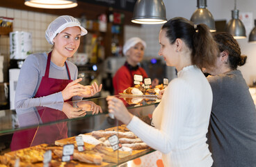Bakery shop girl assistant talks to customer, answers clients questions and recommends fresh products. Employee demonstrates range of baked goods, offers to buy croissant with brie cheese.
