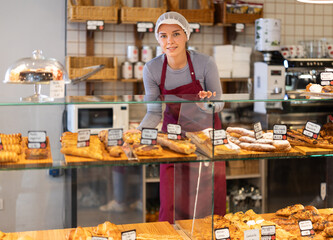 Girl saleswoman works in cafeteria bakery shop, examines assortment and makes decoration of display...
