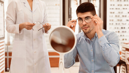 A young man adjusts the frame of glasses while looking in a mirror at an optical store. An...