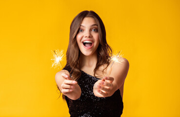 A young woman in a black dress joyfully celebrates with sparklers against a vibrant yellow...