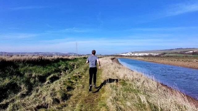 Camera follows man running along grassy trail path next to river on shoreline on spring sunny day (South downs, southease, England)