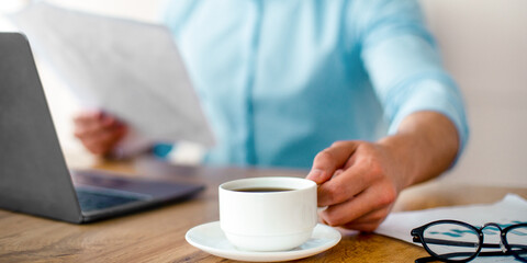A young Middle Eastern businessman sits at his desk, holding a cup of coffee. He reviews important documents, balancing work and lifestyle in a corporate environment during the day.