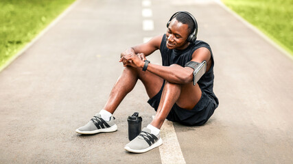 Sporty black guy resting and using his smartwatch, drinking protein, exercising at park, free space
