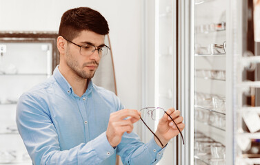 A young man inspects a pair of glasses while browsing in an optical shop. The setting features neatly arranged eyewear displays, creating a stylish environment for customers.