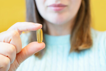 Young woman holding omega 3 capsule on yellow background close-up.