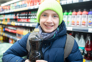 Portrait of 12 year old boy in green hat and blue jacket holding bottle of soda standing in front of shelves with sodas and juices in supermarket.