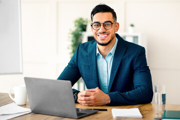 Fototapeta premium A young Arab entrepreneur is smiling while seated at a desk in a contemporary office. He is dressed in a formal suit, engaged in professional work on his laptop with a relaxed demeanor.