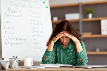 A young Arab woman sits at her desk, visibly stressed while studying lesson plans for remote teaching. She is surrounded by educational materials and a whiteboard filled with notes.