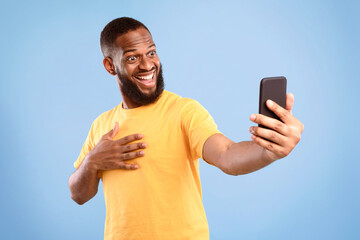 A cheerful African American man captures a selfie with his smartphone, showcasing excitement and...