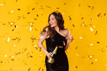 A joyful woman in a black dress holds a champagne bottle and glass, surrounded by golden confetti....