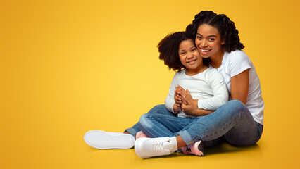 Fototapeta premium Happy Loving Family. Smiling Young African Mother Embracing Her Lovely Little Daughter While Sitting On Floor Over Pink Background Together