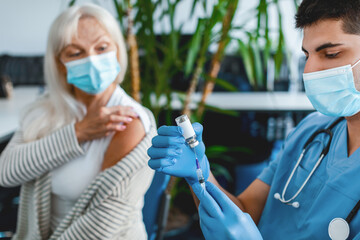 A nurse prepares a vaccine while an elderly woman awaits her immunization. The setting is a modern clinic, emphasizing public health during the flu season amid ongoing pandemic efforts.