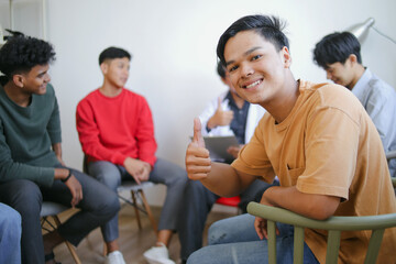 Excited Young Asian Man Giving Thumb Up Against Blurry Group Psychological Counseling Background