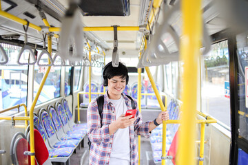 Naklejka premium Young Asian Man Using Smartphone and Listening Music Through Headphones While Commuting by Bus