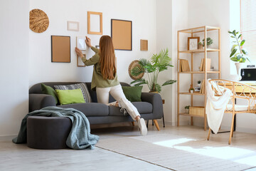 Young woman hanging picture on white wall near sofa in living room