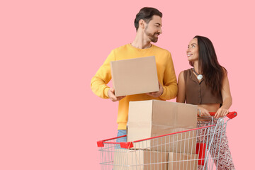 Happy young couple with shopping cart of cardboard boxes on pink background
