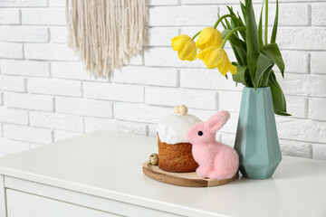 Tulip flowers in vase with Easter cake and rabbit on commode near white brick wall