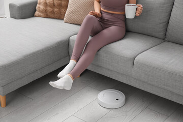 Sporty young woman with cup of tea sitting on sofa and robot vacuum cleaner at home
