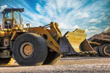 wheel loader at a diamond mine with parked mining trucks, chains on the front wheels to protect tires and for the grip, outdoors sunset with blue sky and sun rays