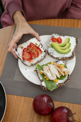 A graceful woman, elegantly dressed, enjoys a refined breakfast featuring a trio of toasts with cream cheese: one topped with fresh tomato slices, another with creamy avocado, and the third with tende