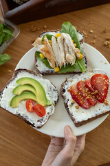 An elegant presentation of three refined toasts, each topped with a smooth layer of cream cheese. One features ripe tomato slices, the second fresh avocado, and the third a tender slice of chicken. Se